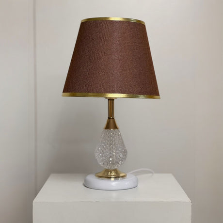 A table lamp with a crystal-textured glass base, gold detailing, and a brown fabric lampshade with gold trim, set against a plain light-colored wall.