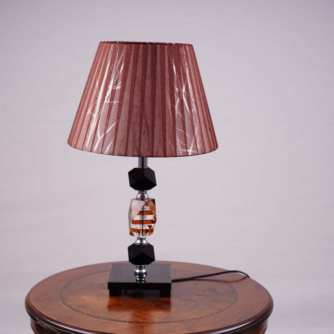 An elegant brown table lamp featuring a classic white lampshade and a sleek black-and-transparent glass base, placed on a wooden table. Equipped with an E27 LED light, it radiates a warm and sophisticated glow, enhancing the ambiance with a touch of refinement.