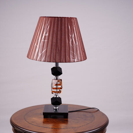 An elegant brown table lamp featuring a classic white lampshade and a sleek black-and-transparent glass base, placed on a wooden table. Equipped with an E27 LED light, it radiates a warm and sophisticated glow, enhancing the ambiance with a touch of refinement.