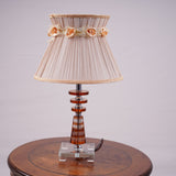 Elegant table lamp with a beige pleated lampshade, adorned with delicate fabric roses and gold trim. The base is crafted from a combination of transparent and amber-colored glass with a geometric design, supported by a sturdy glass platform. Equipped with an E27 LED light, it emits a warm and sophisticated glow. Placed on a round wooden table.