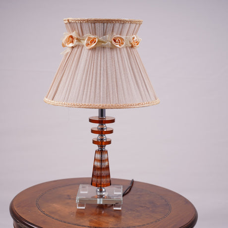 Elegant table lamp with a beige pleated lampshade, adorned with delicate fabric roses and gold trim. The base is crafted from a combination of transparent and amber-colored glass with a geometric design, supported by a sturdy glass platform. Equipped with an E27 LED light, it emits a warm and sophisticated glow. Placed on a round wooden table.