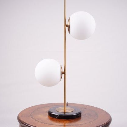 A modern gold table lamp with two frosted glass globes on a black circular base, set on a wooden table.
