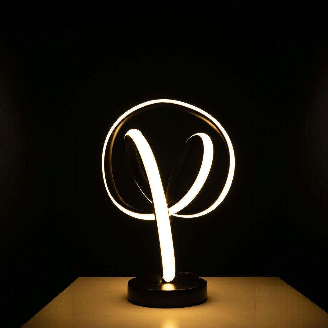 The LED table lamp has a black base with intertwined infinity-loop shapes. The loops are lit up with soft white light, creating a glowing effect. it sits  on  a wooden table with dark background