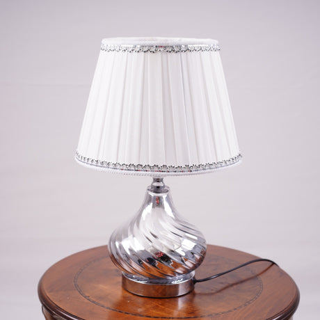 An elegant table lamp with a chic twisted silver base and a classic white lampshade. Equipped with an E27 LED light, it casts a warm and sophisticated glow.