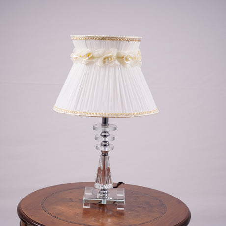 Elegant table lamp with a white pleated lampshade, adorned with delicate fabric roses and gold trim. The base is crafted from a combination of transparent and amber-colored glass with a geometric design, supported by a sturdy glass platform. Equipped with an E27 LED light, it emits a warm and sophisticated glow. Placed on a round wooden table.
