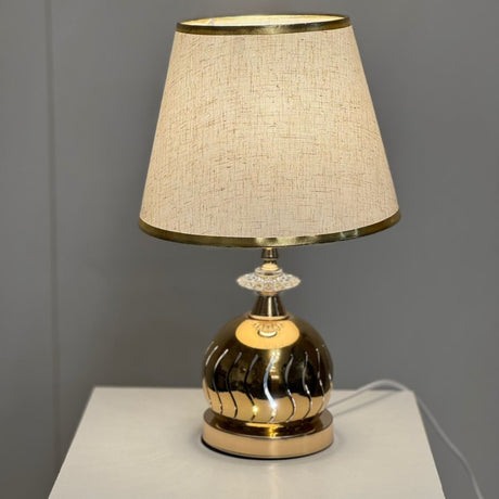 Gold and beige table lamp with a round metallic gold base and a beige fabric lampshade with delicate gold trim, placed on a white table.
