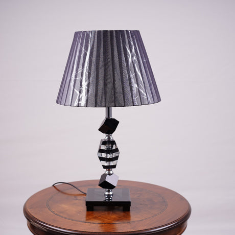 An elegant black table lamp featuring a classic white lampshade and a sleek black-and-transparent glass base, placed on a wooden table. Equipped with an E27 LED light, it radiates a warm and sophisticated glow, enhancing the ambiance with a touch of refinement.