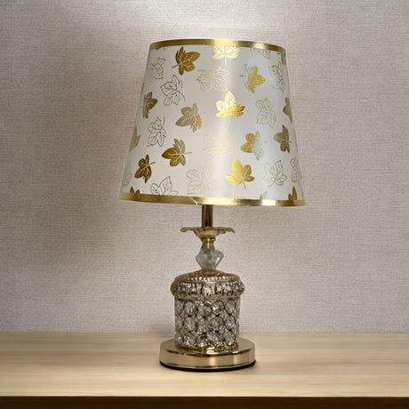 A luxurious table lamp with an ornate gold and glass base, and a white lampshade with gold leaf patterns, positioned on a wooden table against a neutral wall.