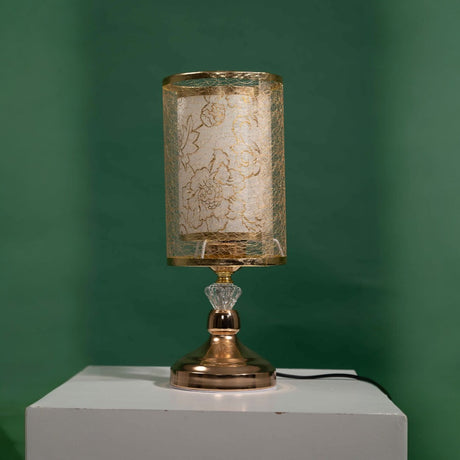 Gold table lamp with a cylindrical transparent shade decorated with a leaf-like etched design, equipped with a led lamp E27, placed on a white table and green background.