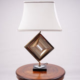 A modern table lamp with a unique geometric design, featuring a gold metal base in the shape of a rotated square with intricate latticework. The lamp is placed on a round wooden table and has a white fabric shade with a slightly curved design, accented by two brown lines along the top and bottom edges, adding elegance to the overall look.