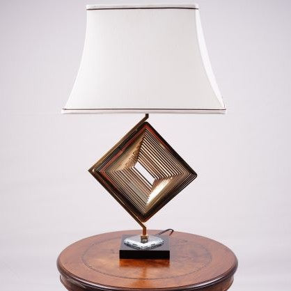 A modern table lamp with a unique geometric design, featuring a gold metal base in the shape of a rotated square with intricate latticework. The lamp is placed on a round wooden table and has a white fabric shade with a slightly curved design, accented by two brown lines along the top and bottom edges, adding elegance to the overall look.