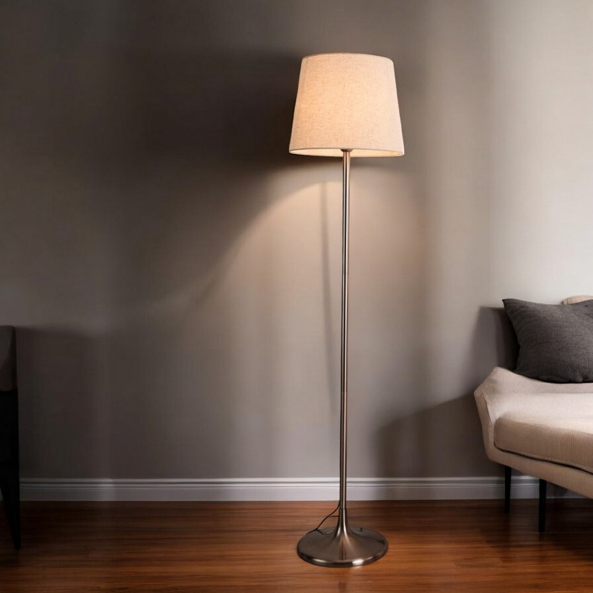 A simple and elegant floor lamp with a slim metallic base and a beige lampshade, standing against a grey wall next to a beige armchair.