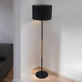 A tall, slim black floor lamp with a black cylindrical lampshade and wooden accents, standing in a modern living space.