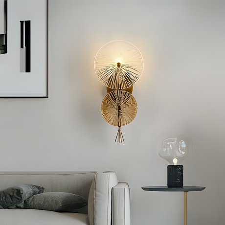 Modern decorative wall light with two fan-like glass discs on a gold mount, installed above a neutral-toned sofa and beside a sleek black side table with a translucent glass lamp.