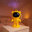 Yellow astronaut-like night light on a bedside table with a starry sky projection in the background.