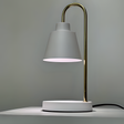 The desk lamp features a sleek and minimalist design with a white lampshade resting on a slender gold stand. The stand curves from a circular white base.  Positioned against a neutral background.