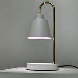 The desk lamp features a sleek and minimalist design with a white lampshade resting on a slender gold stand. The stand curves from a circular white base.  Positioned against a neutral background.