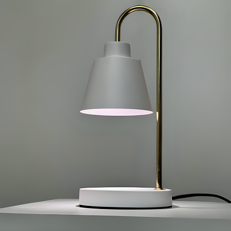 The desk lamp features a sleek and minimalist design with a white lampshade resting on a slender gold stand. The stand curves from a circular white base.  Positioned against a neutral background.