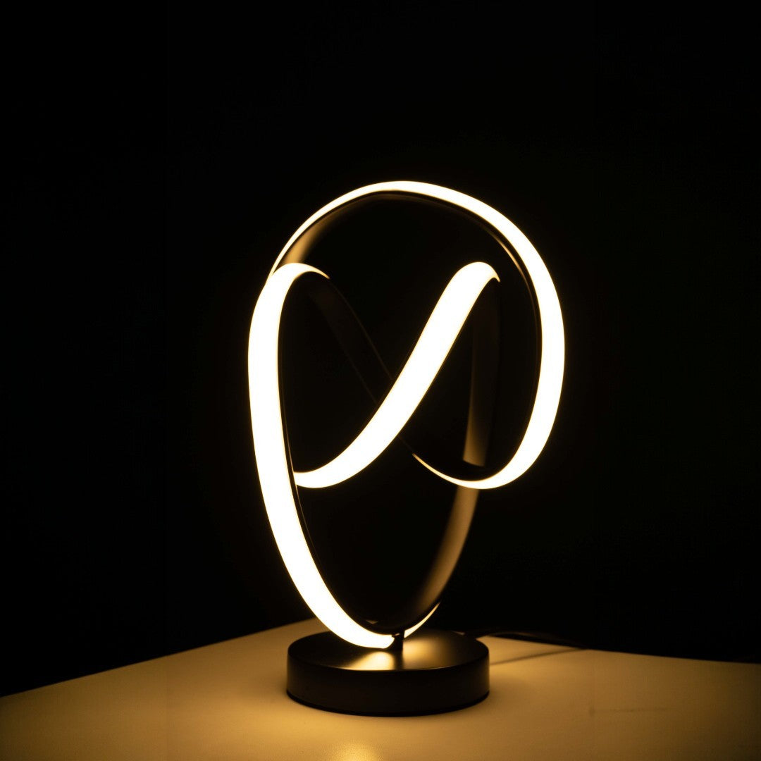 The LED table lamp has a black base with intertwined infinity-loop shapes. The loops are lit up with soft white light, creating a glowing effect. it sits  on  a wooden table with dark background