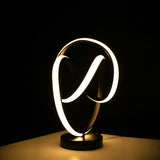 The LED table lamp has a black base with intertwined infinity-loop shapes. The loops are lit up with soft white light, creating a glowing effect. it sits  on  a wooden table with dark background