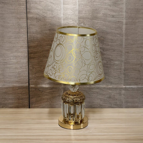 A table lamp with a decorative gold and transparent glass base, and a beige lampshade adorned with gold circular patterns, placed on a wooden surface with a textured wall in the background.