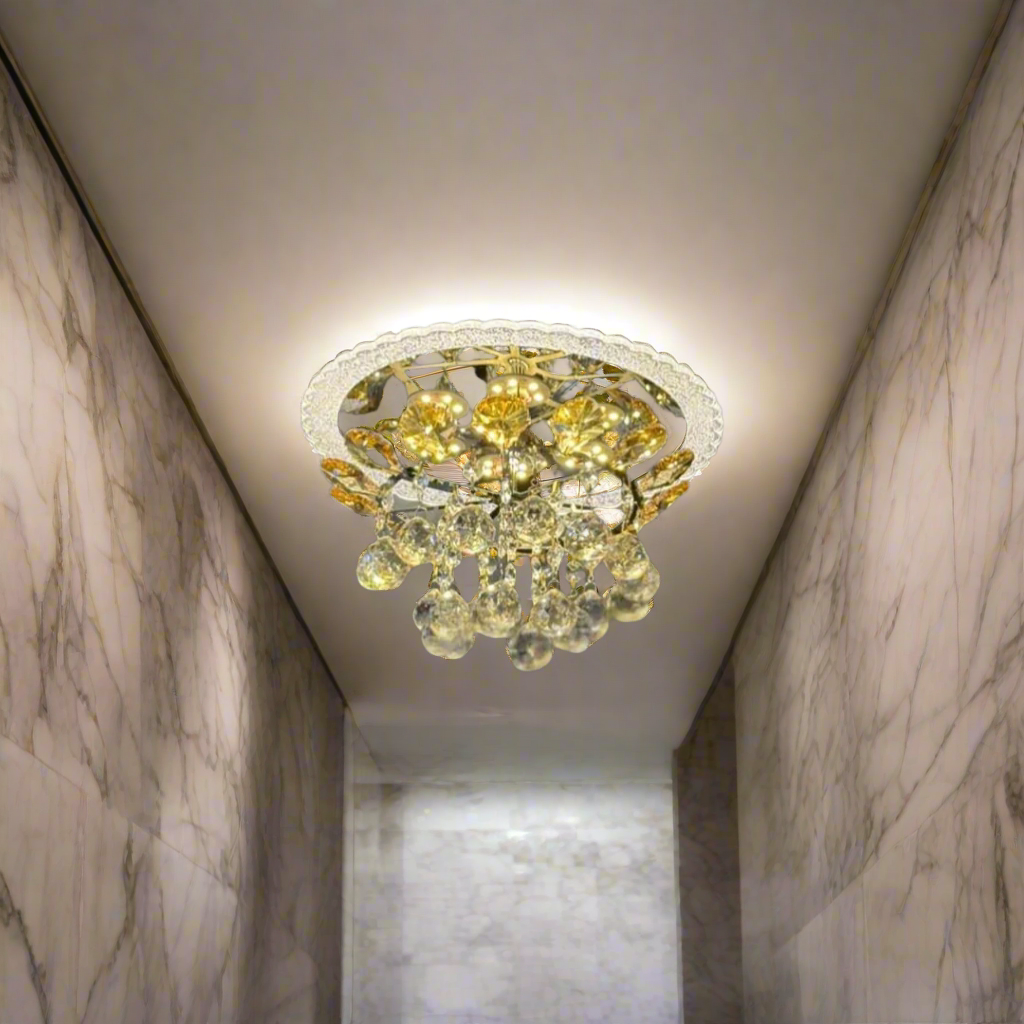 A compact, ceiling-mounted chandelier featuring a gold-toned metal frame and a cluster of teardrop-shaped crystal pendants. The fixture casts warm LED lighting, creating a decorative sunburst reflection on the ceiling. 
