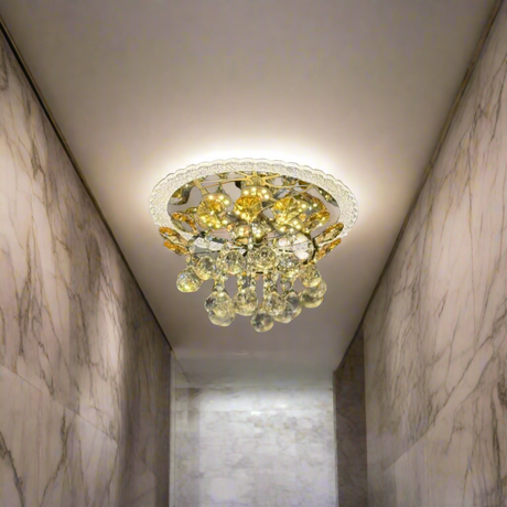 A compact, ceiling-mounted chandelier featuring a gold-toned metal frame and a cluster of teardrop-shaped crystal pendants. The fixture casts warm LED lighting, creating a decorative sunburst reflection on the ceiling. 