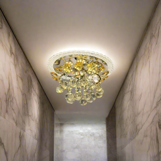 A compact, ceiling-mounted chandelier featuring a gold-toned metal frame and a cluster of teardrop-shaped crystal pendants. The fixture casts warm LED lighting, creating a decorative sunburst reflection on the ceiling. 