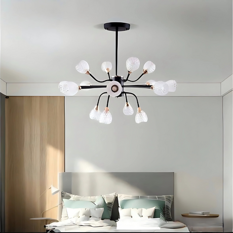 A modern bedroom featuring a stylish black and gold chandelier with multiple curved arms, each ending in a frosted, petal-shaped glass light. The room has minimalist decor with a gray upholstered headboard, patterned pillows, and soft neutral tones.