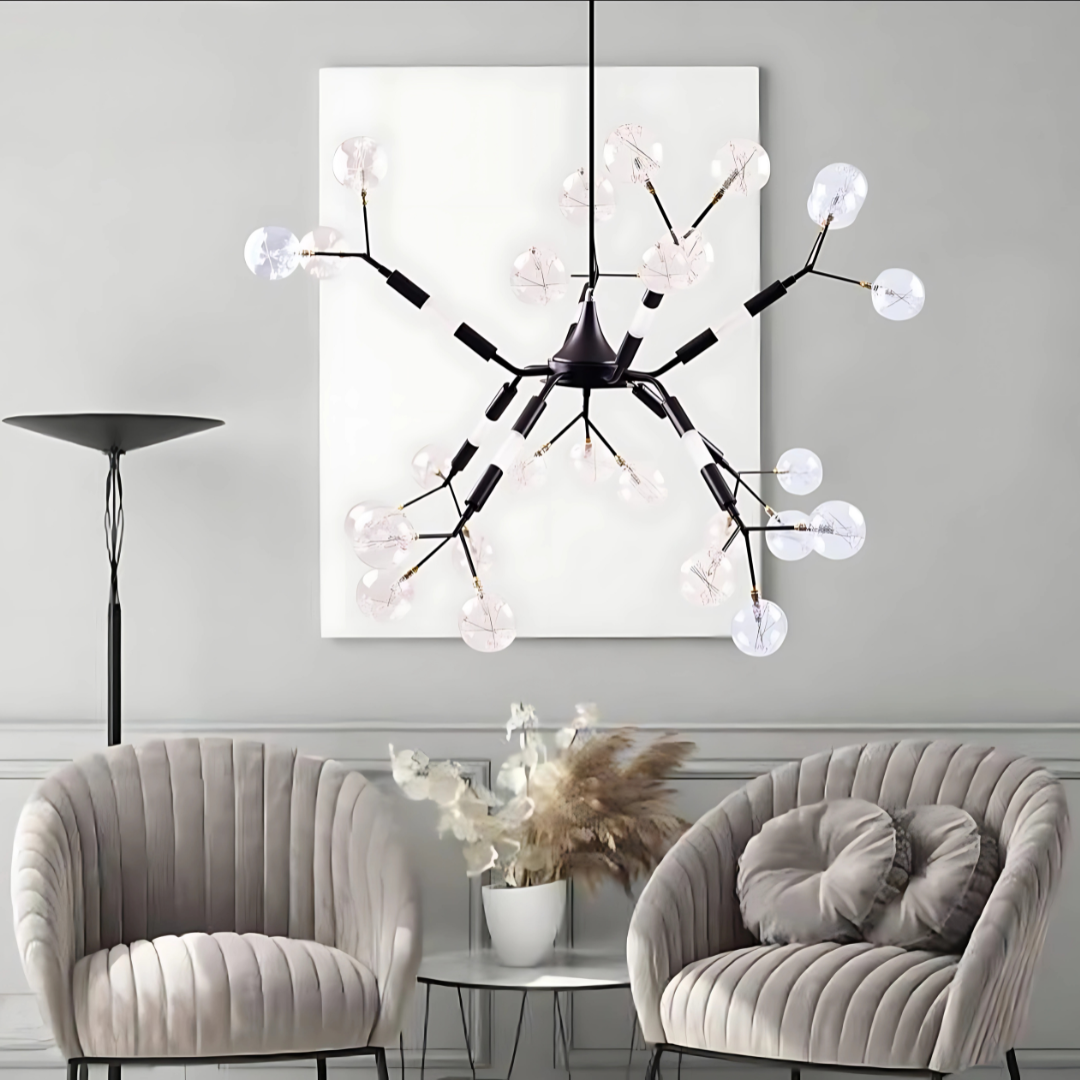 A modern living room with a decorative black ceiling light featuring multiple curved arms, each ending in a clear, petal-shaped glass bulb. The room includes two plush grey chairs, a glass coffee table with a vase of dried flowers, and a tall floor lamp. 