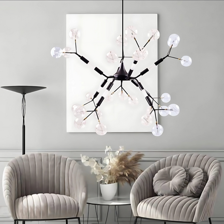 A modern living room with a decorative black ceiling light featuring multiple curved arms, each ending in a clear, petal-shaped glass bulb. The room includes two plush grey chairs, a glass coffee table with a vase of dried flowers, and a tall floor lamp. 