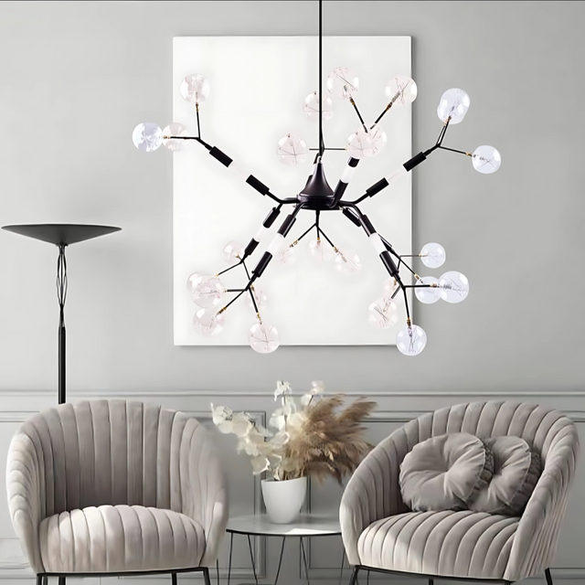 A modern living room with a decorative black ceiling light featuring multiple curved arms, each ending in a clear, petal-shaped glass bulb. The room includes two plush grey chairs, a glass coffee table with a vase of dried flowers, and a tall floor lamp. 
