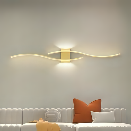 An artistic gold wall light with curving, wave-like LED elements, mounted above a sofa with orange and white cushions.