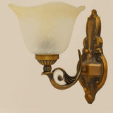 A classic gold wall sconce with a curved arm and frosted glass shade, featuring intricate detailing on the mounting plate.
