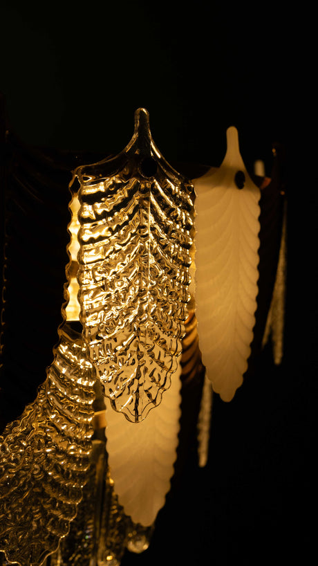  A close-up view of a luxurious chandelier featuring gold and white leaf-shaped plexyglass ornaments, illuminated to create a warm and opulent glow.