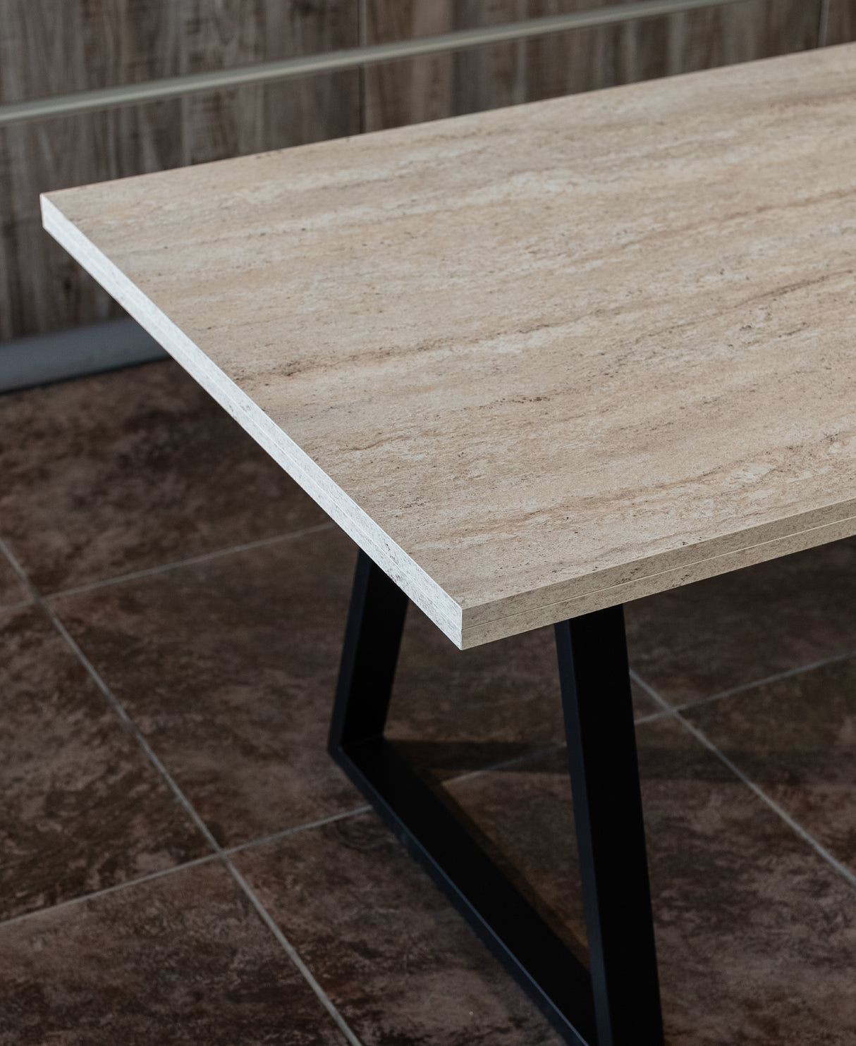 Close-up view of a rectangular dining table with a natural stone-look melamine wood top and black metal V-frame legs, highlighting the texture and edge detail of the tabletop.

