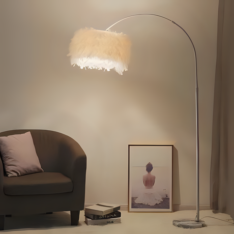  A curved floor lamp with a white feathered lampshade and a sleek metallic stand, placed in a cozy corner beside a brown armchair and a framed artwork on the floor.
