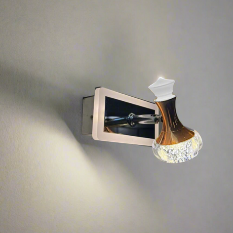 A modern wall-mounted LED light fixture with a metallic rectangular base and an ornate, bottle-shaped glass lampshade that has a golden-brown upper section and a textured, translucent lower section. The fixture is mounted on a plain, light-colored wall and emits a soft, downward glow.