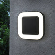 A modern outdoor wall light with a square black center and glowing white border, mounted on a dark exterior wall.