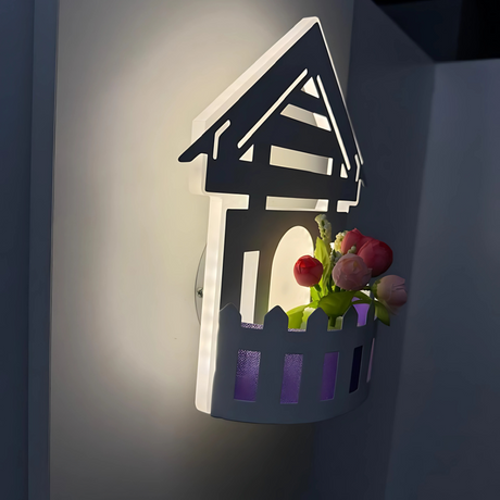 Wall-mounted LED light fixture designed in the shape of a white house , featuring a cork-style built-in compartment holding artificial pink and red tulip flowers. The light has a whimsical, cottage-inspired aesthetic and is mounted on a plain white wall.
