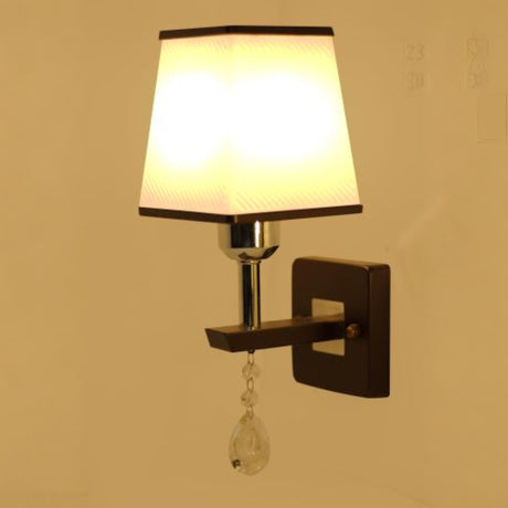 A brown and silver wall light with a rectangular fabric shade and a hanging crystal ornament, adding a touch of elegance turned on, casting warm light.
