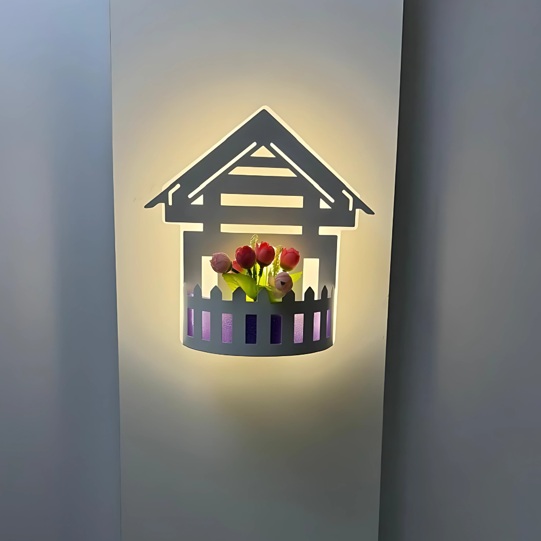 Wall-mounted LED light fixture designed in the shape of a white house , featuring a cork-style built-in compartment holding artificial pink and red tulip flowers. The light has a whimsical, cottage-inspired aesthetic and is mounted on a plain white wall.
