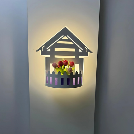 Wall-mounted LED light fixture designed in the shape of a white house , featuring a cork-style built-in compartment holding artificial pink and red tulip flowers. The light has a whimsical, cottage-inspired aesthetic and is mounted on a plain white wall.
