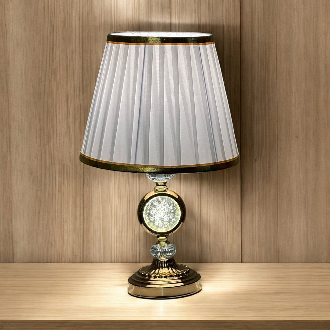 Elegant gold table lamp with a pleated white shade with a gold trim and a crystal accent in the center, placed on a wooden surface against a wood-paneled background.