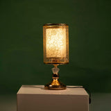 Gold table lamp with a cylindrical transparent shade decorated with a leaf-like etched design, equipped with a led lamp E27, placed on a white table and green background.
