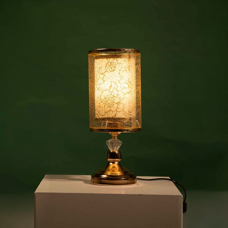 Gold table lamp with a cylindrical transparent shade decorated with a leaf-like etched design, equipped with a led lamp E27, placed on a white table and green background.
