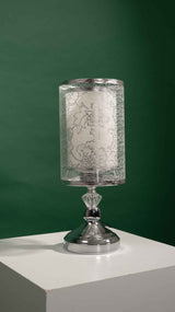 Silver table lamp with a cylindrical transparent shade decorated with a leaf-like etched design, placed on a white table and green background.
