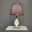A table lamp with a white textured glass base, gold accents, and a brown fabric lampshade with delicate gold trim, placed on a white surface against a neutral grey background.
