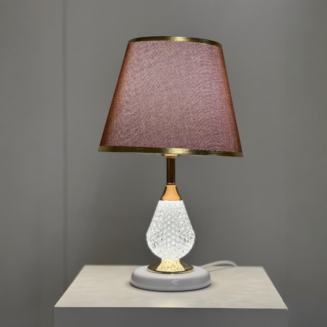A table lamp with a white textured glass base, gold accents, and a brown fabric lampshade with delicate gold trim, placed on a white surface against a neutral grey background.