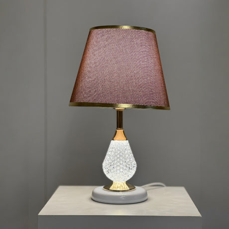 A table lamp with a white textured glass base, gold accents, and a brown fabric lampshade with delicate gold trim, placed on a white surface against a neutral grey background.